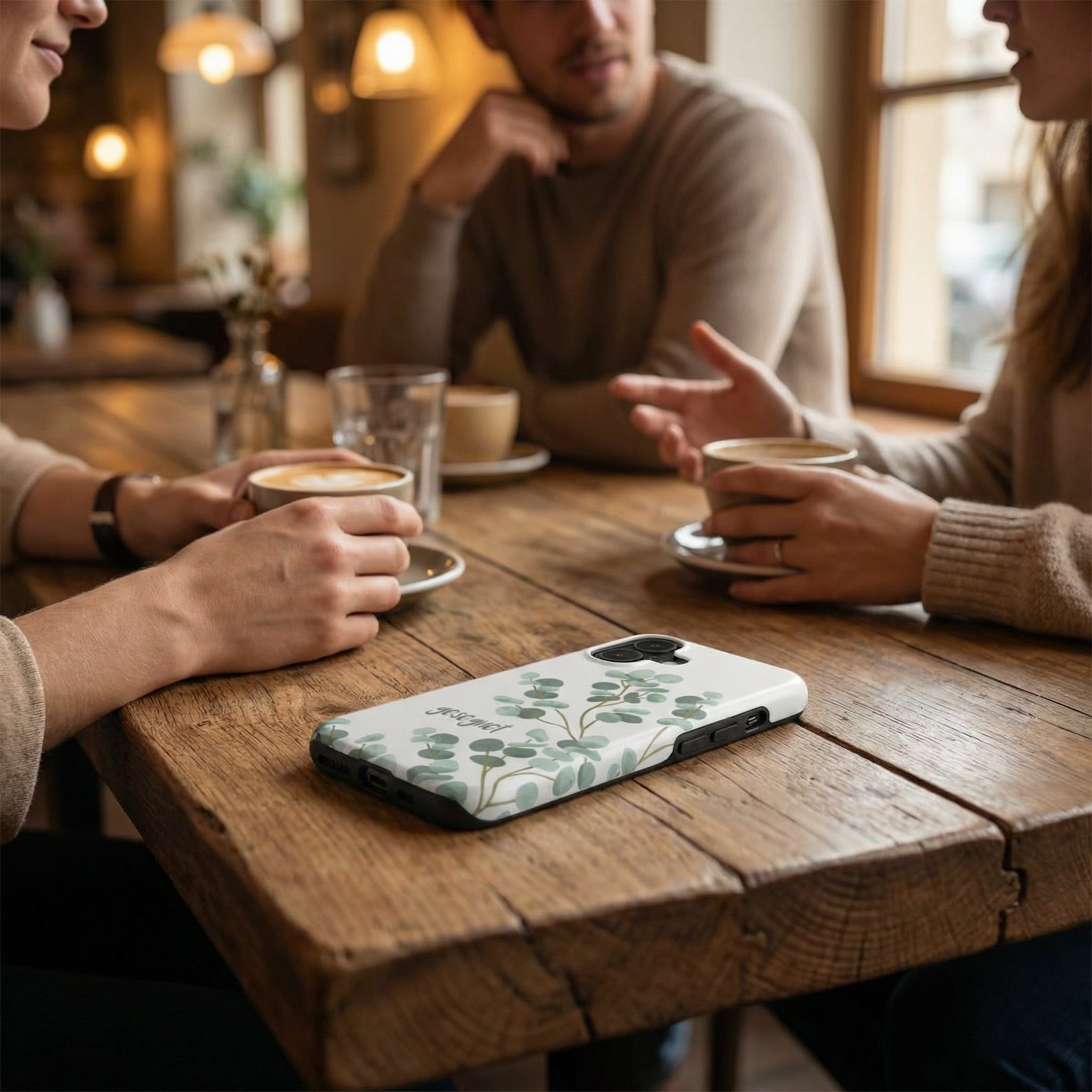 Handyhülle mit Motiv GESEGNET in Weiß mit grünem Eukalyptus-Design liegt auf Holztisch im Café – Lifestyle