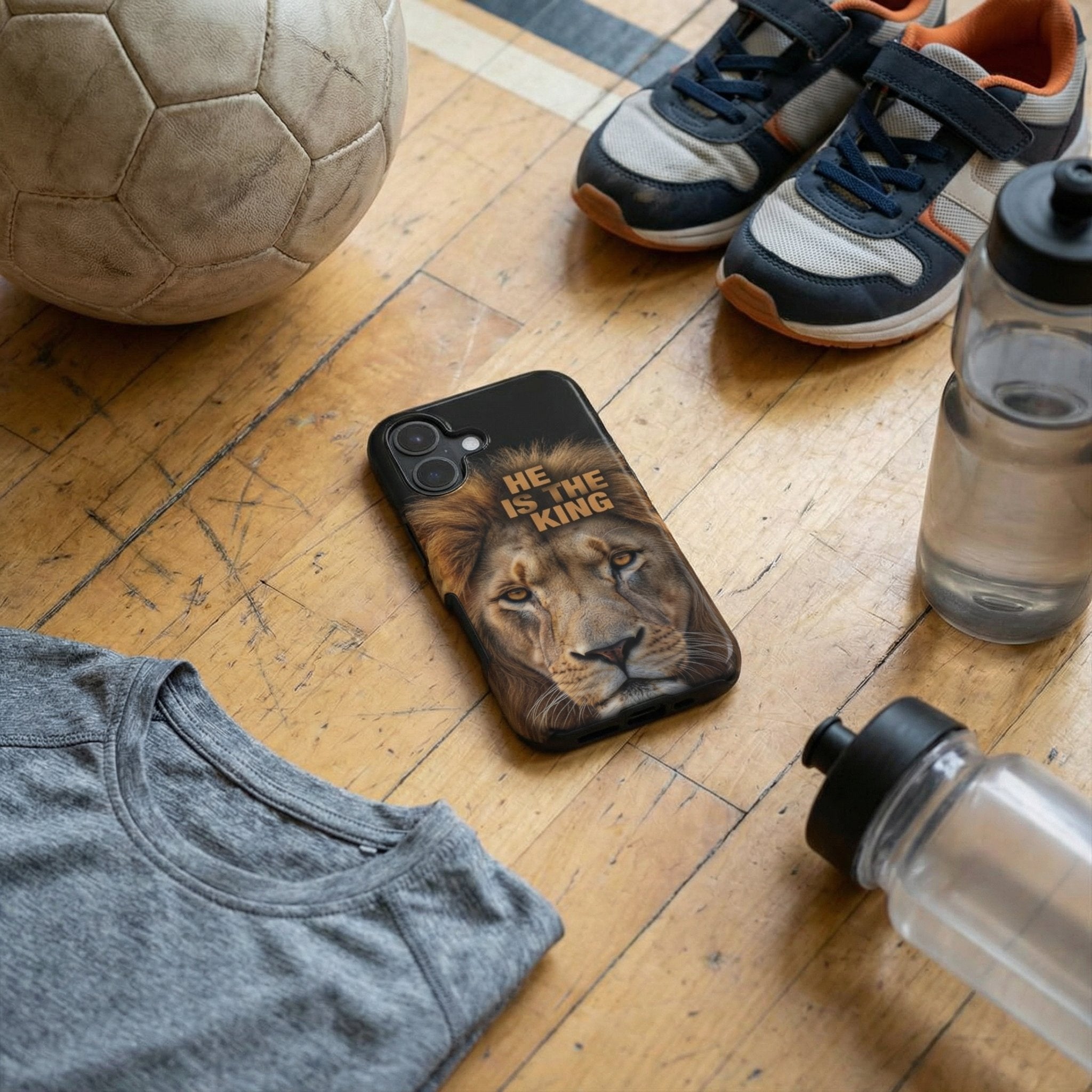 Handyhülle Glaube mit HE IS THE KING und Löwen-Design auf Holzboden neben Fußball und Sportschuhen – Lifestyle